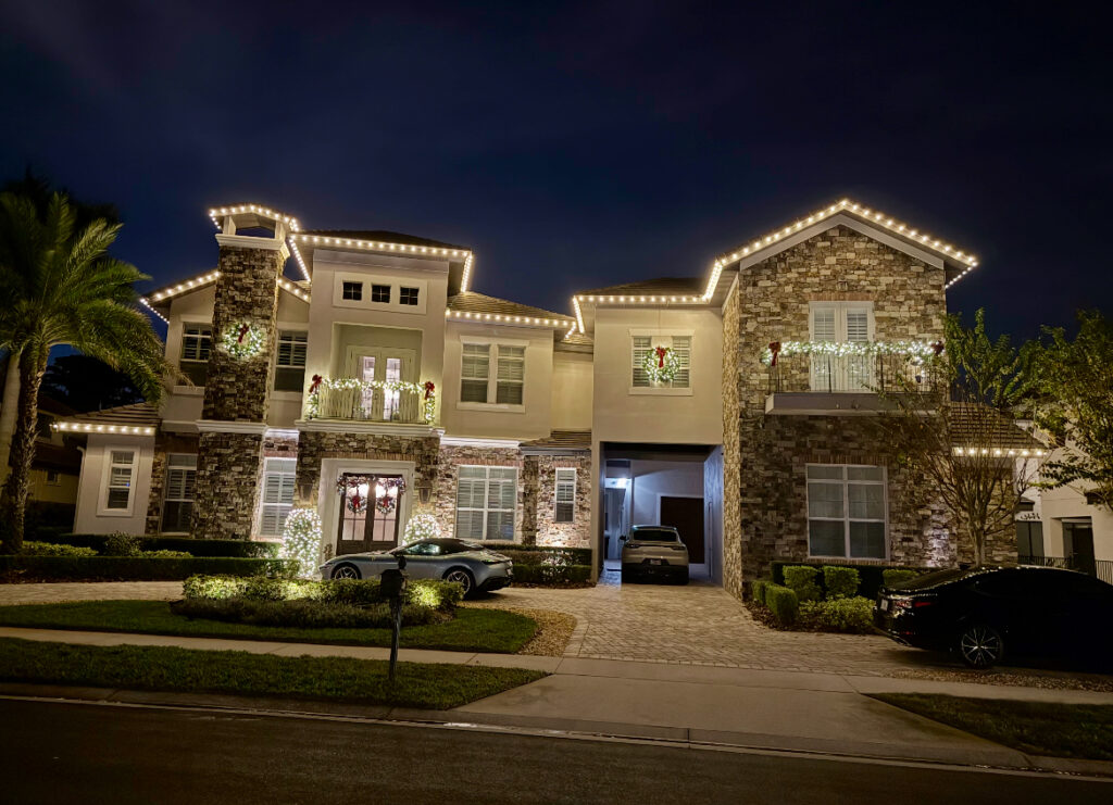 Two-story stone and stucco home with warm white roofline lights, lit balcony garland and window wreaths
