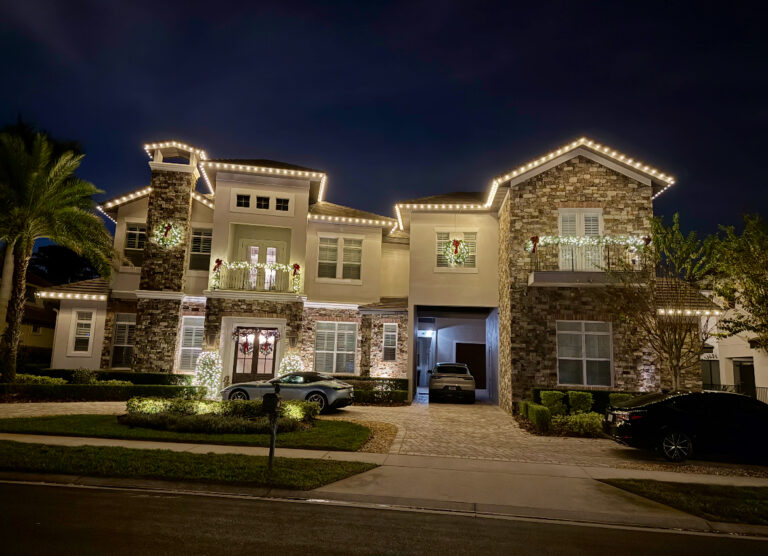 Two-story stone and stucco home with warm white roofline lights and lit balcony garland