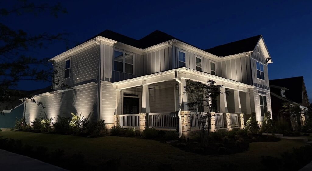 Two-story home with a deep front porch lit by warm landscape uplighting